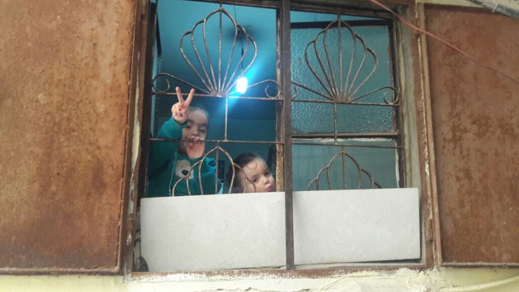 طفلة تقف هي وشقيقتها على نافذة منزلهما رافعة علامة النصر في مخيم برج البراجنة للاجئين الفلسطينيين في ضاحية بيروت الجنوبية (بعدسة سامر العلي)
