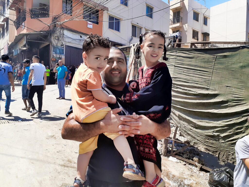 أب وطفليه يبتسمون لعدسة “شبابيك” في مخيم نهر البارد للاجئين الفلسطينيين في مدينة طرابلس شمال لبنان (بعدسة عدنان الحمد)