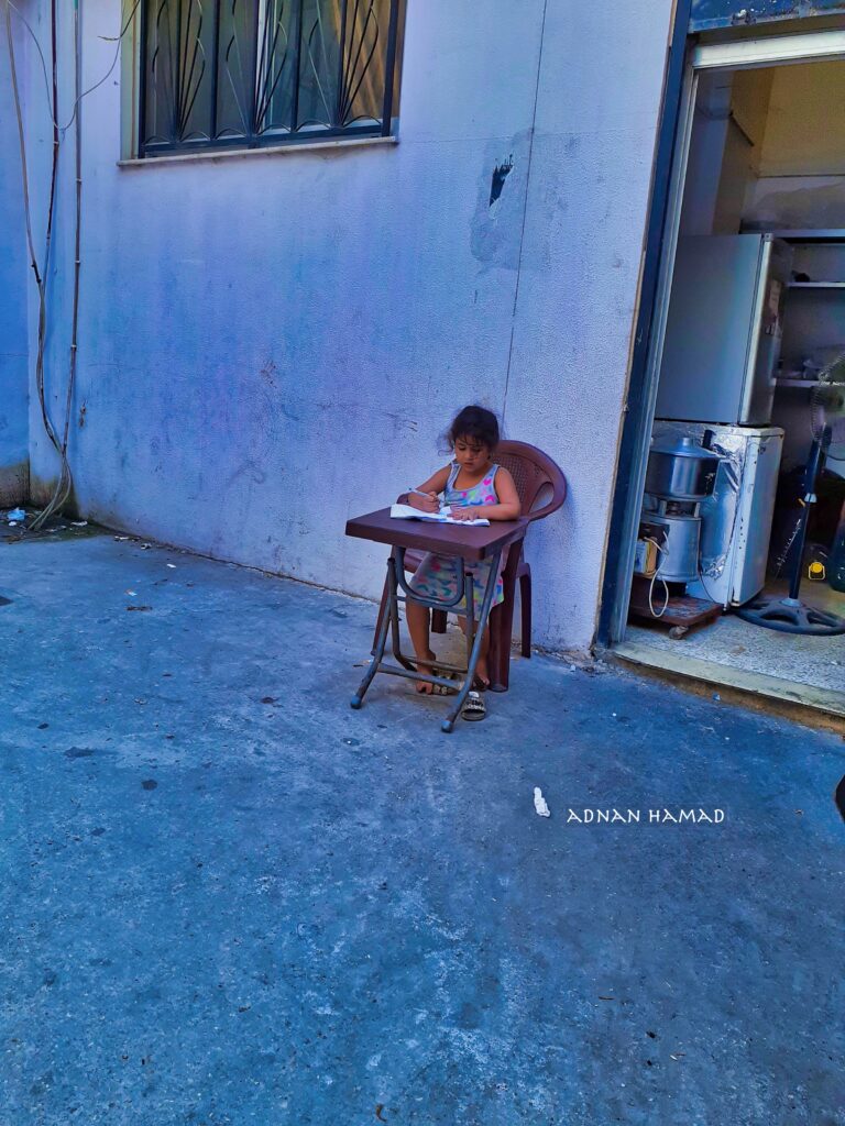 طفلة تقوم بمراجعة دروسها على حافة الطريق بالقرب من منزلها في مخيم نهر البارد للاجئين الفلسطينيين في مدينة طرابلس، شمال لبنان (بعدسة عدنان الحمد)