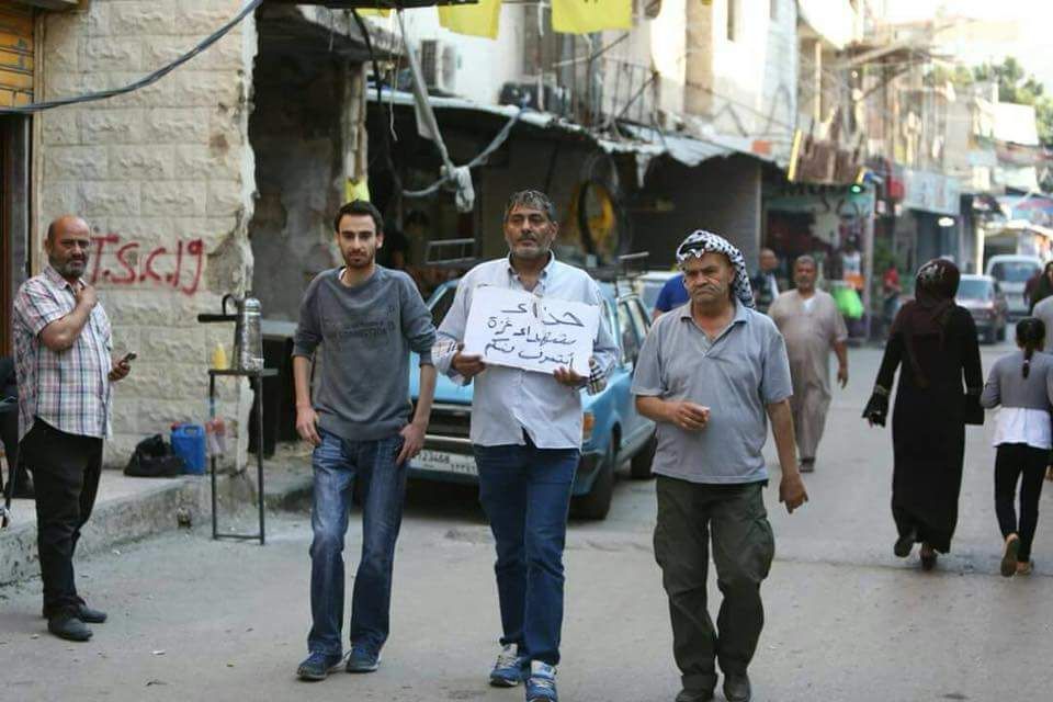ابراهيم ميعاري الملقب ب “ابو الحن” يجول في شوارع مخيم عين الحلوة، في صيدا، جنوب لبنان، رافعا يافطة كُتب عليها “حذاء شهداء غزة أشرف منكم” (بعدسة رشا حيدر)